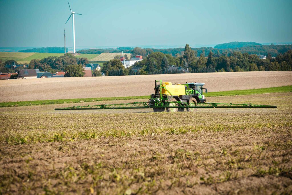 Grüner Dammann Profi Class Feldspritze auf einem Acker mit Windrad im Hintergrund unter blauem Himmel.