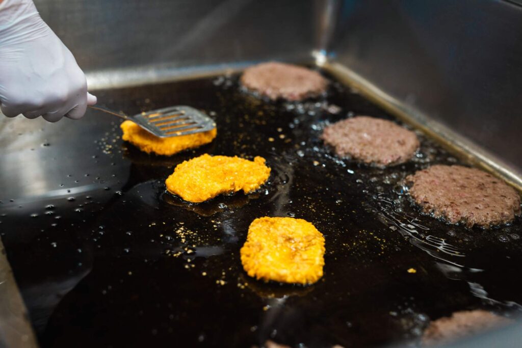 Nahaufnahme des Bratens von vegetarischen/veganen Kartoffel- Patties und braunen Fleisch-Patties auf einer heißen, stark geölten Grillplatte. Eine Hand mit weißem Handschuh wendet einen Patty mit einem Pfannenwender.