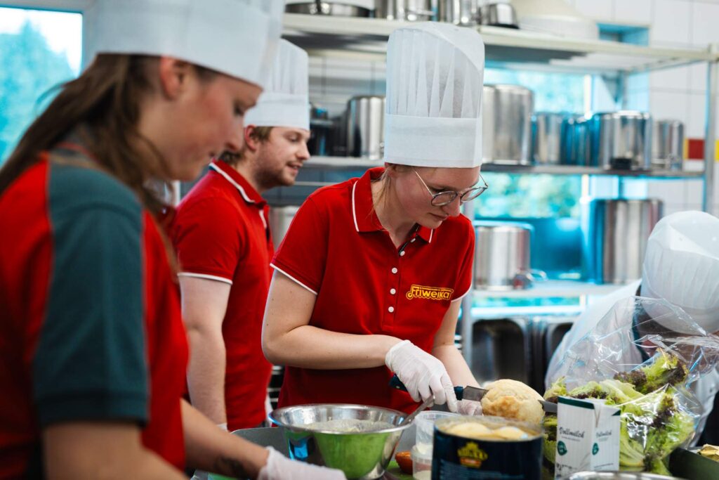 Drei Köche in roten Friweika-Shirts und weißen Kochmützen bei der Arbeit in der Küche. Die vordere Köchin mit Brille schneidet mit Handschuhen ein Brötchen zu.