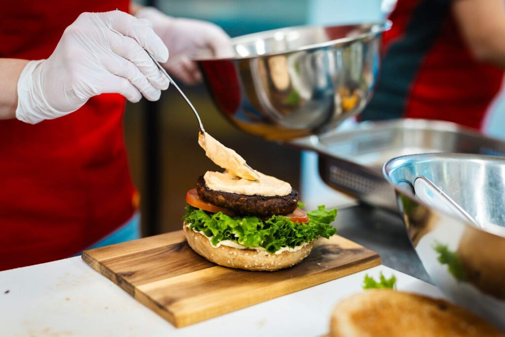 Detailaufnahme der Zubereitung eines Burgers: Eine Hand mit Handschuh löffelt Soße auf ein Patty, das auf Salat, Tomate und einem Brötchen auf einem kleinen Holzbrett liegt.
