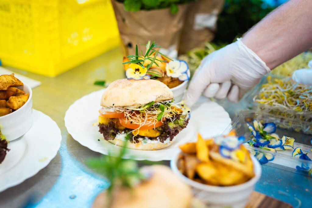 Nahaufnahme eines kunstvoll angerichteten Burgers auf einem weißen Teller, garniert mit Sprossen, Rosmarinzweigen und essbaren Blüten. Eine Hand mit weißem Handschuh serviert das Gericht.