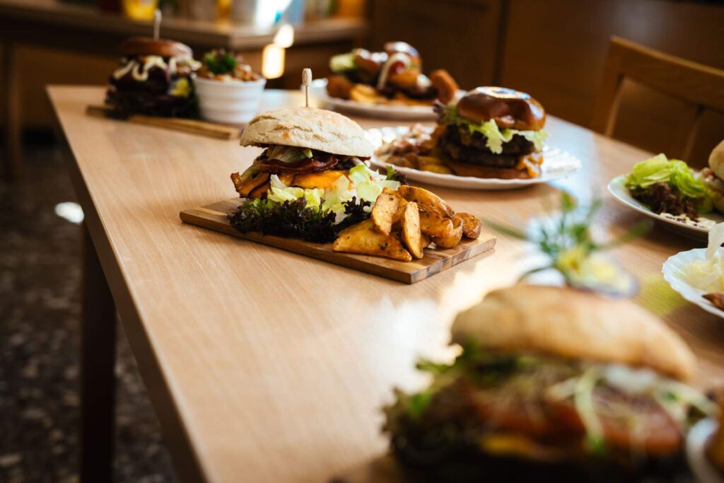Eine Reihe von fertigen, appetitlich angerichteten Burgern auf Holzbrettern und Tellern, die auf einem hellen Holztisch präsentiert werden, mit Kartoffelspalten oder Pommes als Beilage.