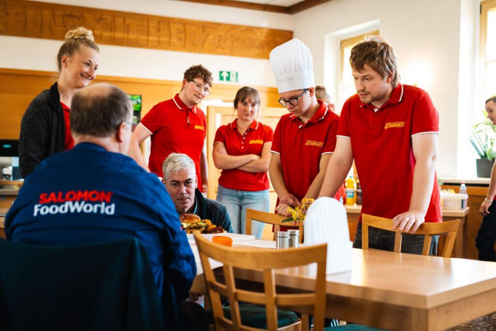Eine Gruppe von Köchen in roten Friweika-Shirts und ein Koch mit Mütze präsentieren ihr Gericht am Holztisch einem Richter, der ein blaues 'SALOMON FoodWorld'-Shirt trägt, während sie das Ergebnis des Burger-Wettbewerbs besprechen.