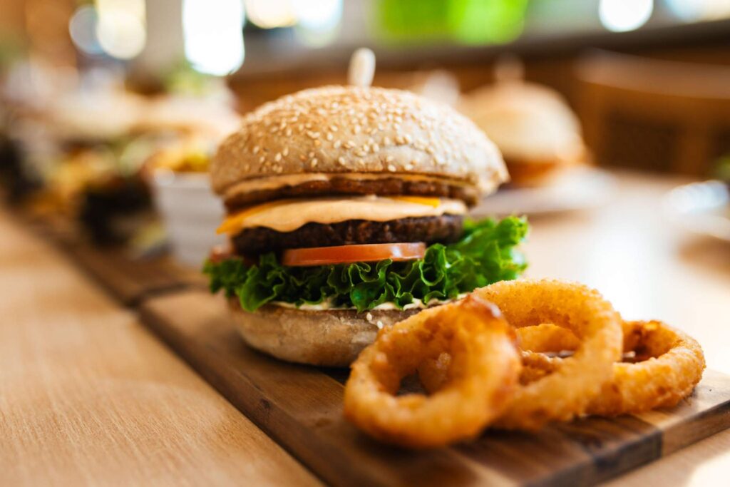 Nahaufnahme eines fertigen Burgers mit Käse, Salat und Tomate auf einem Holzbrett, begleitet von drei goldbraunen, frittierten Zwiebelringen.
