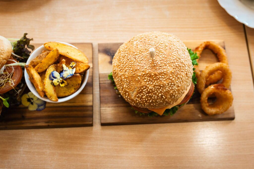 Overhead-Aufnahme von zwei Burger-Gerichten auf Holzbrettern: Links eine Schale Kartoffelspalten (Wedges) mit essbaren Blüten und rechts ein Burger mit Sesambun und drei knusprigen Zwiebelringen.