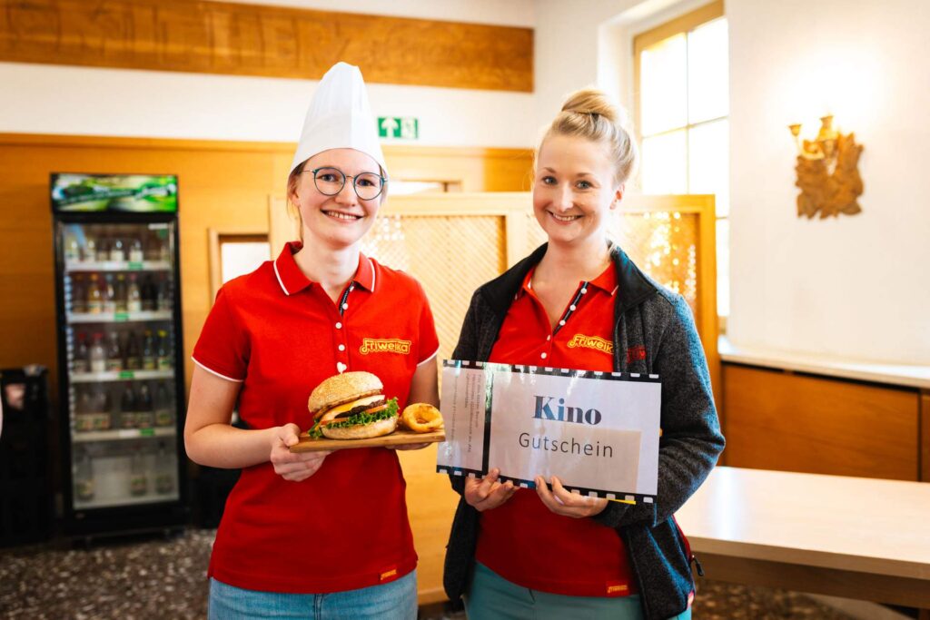 Zwei lächelnde Frauen in Friweika-Shirts präsentieren die Gewinne des Burger-Wettbewerbs. Die Gewinnerin in Kochmütze hält einen Burger auf einem Holzbrett, die andere Frau hält einen 'Kino Gutschein'.