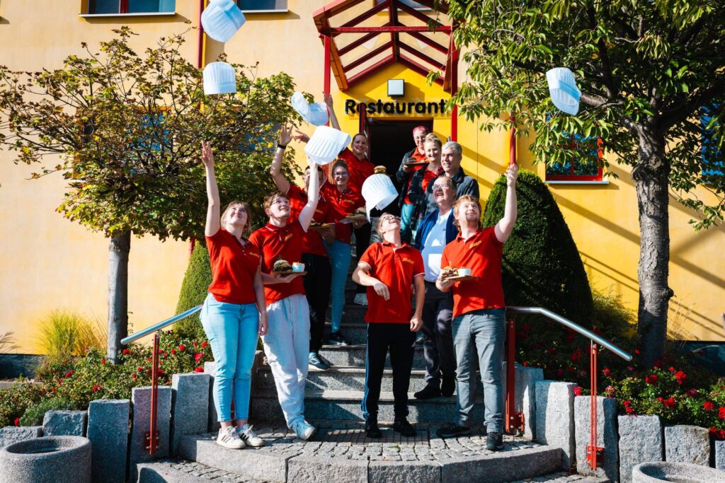 Eine Gruppe von Köchen in roten Friweika-Shirts und Juroren feiert den Abschluss des Burger-Wettbewerbs, indem sie ihre weißen Kochmützen in die Luft werfen. Die Gruppe steht lächelnd auf einer Steintreppe vor einem gelben Gebäude mit einem 'Restaurant'-Schild.