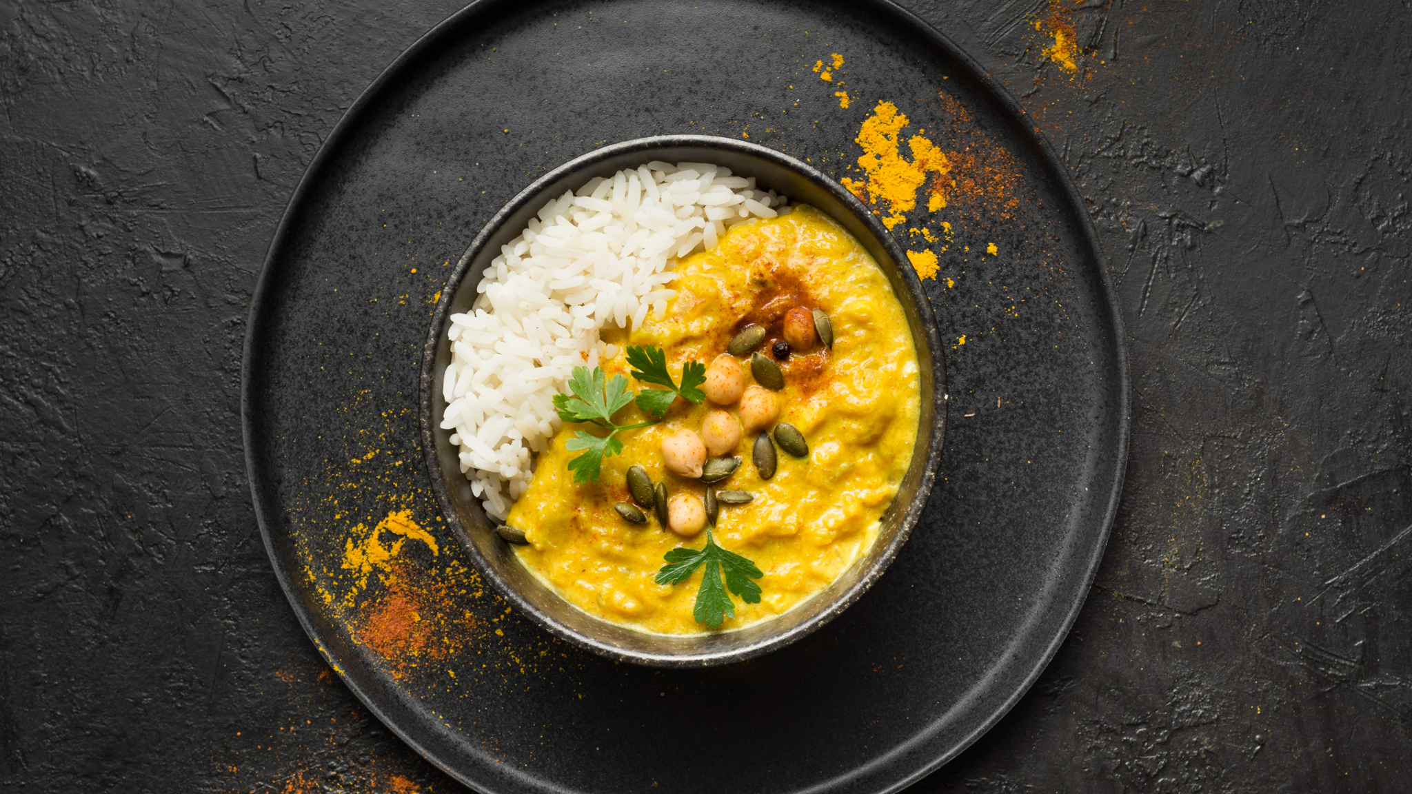 Gelbes Kartoffelcurry mit Reis in einer schwarzen Schale, aufm schwarzem Teller und Tisch angerichtet, getoppt mit Petersilienblättern und ein paar Kernen.