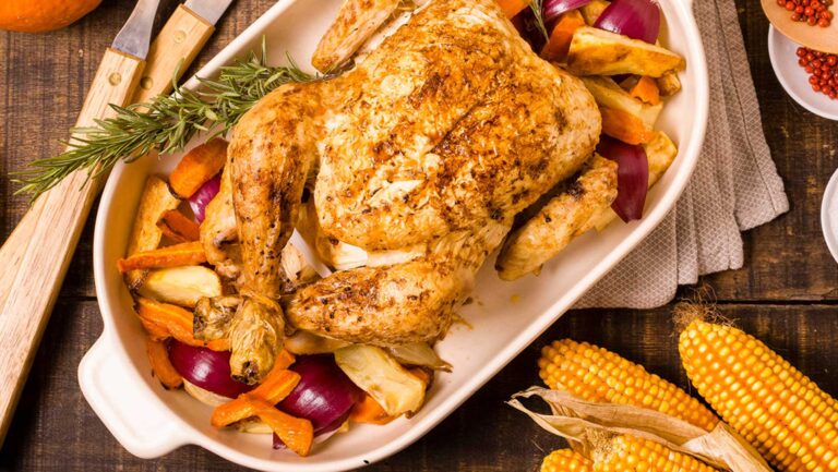 Herbstpfanne aus dem Ofen: Goldbraun gebratenes ganzes Hähnchen umgeben von Kürbis, Zwiebeln und Wurzelgemüse in einer weißen Auflaufform, dekoriert mit Maiskolben, Mandeln und Gewürzen auf rustikalem Holzhintergrund.