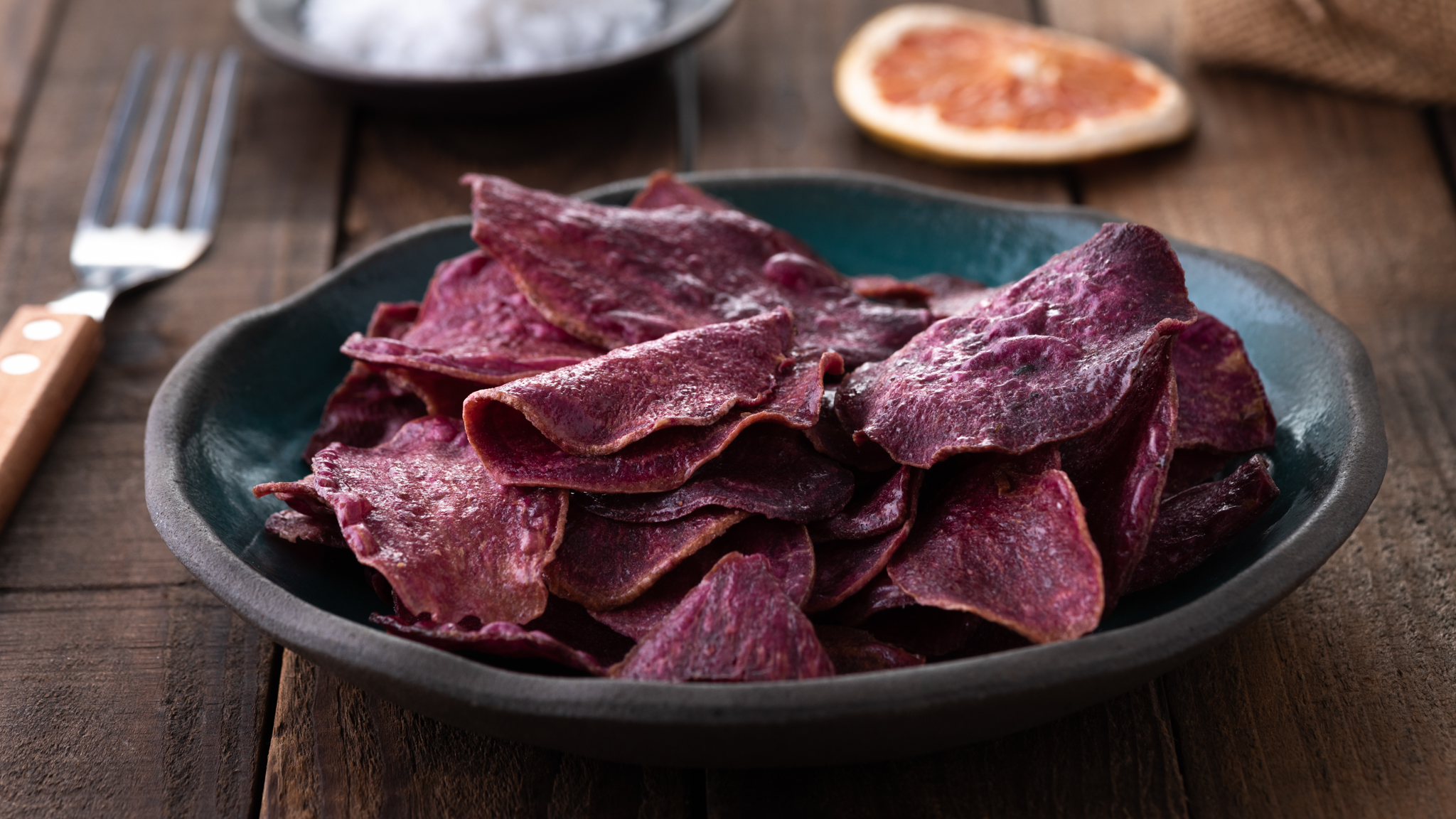 Nahaufnahme von lila Kartoffel-Chips. Die Chips sind in einer dunkelblauen Keramikschale angerichtet und liegen auf einem rustikalen Holztisch. Im unscharfen Hintergrund sind links eine Gabel und eine kleine Schale mit Salz sowie rechts eine getrocknete Zitrusfruchtscheibe und ein Stück Jute zu erkennen.