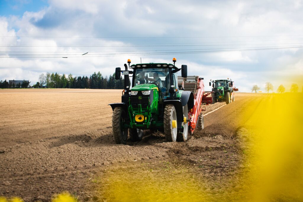 Zwei grüne John Deere Traktoren mit roten Erntemaschinen bei der Feldarbeit auf einem braunen Acker unter bewölktem Himmel.