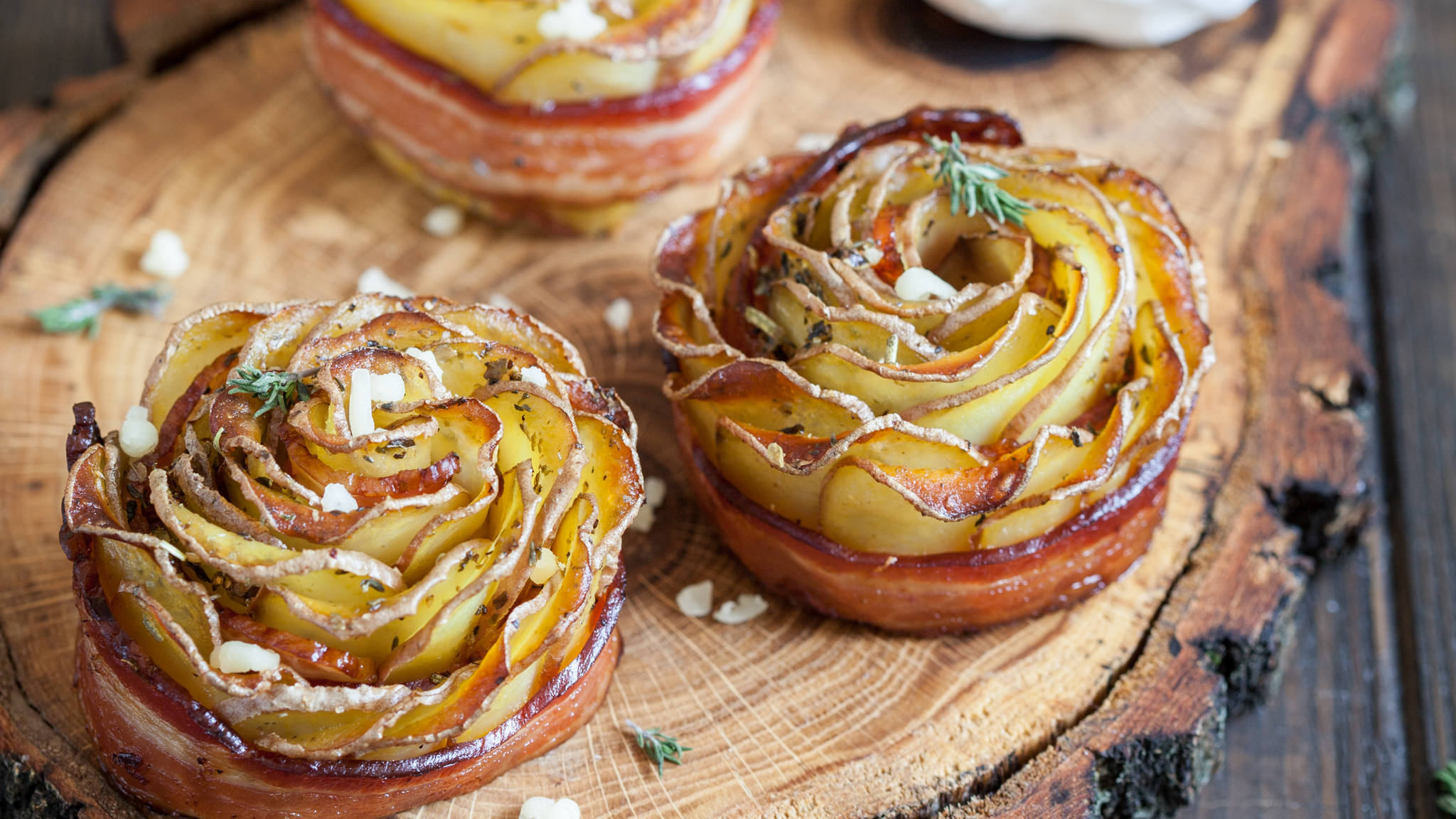 Drei goldbraun gebackene Kartoffel-Rosen mit Speckrand auf einem Holzschneidebrett, garniert mit Kräutern und Meersalz. Im Hintergrund eine Schale mit Dip.