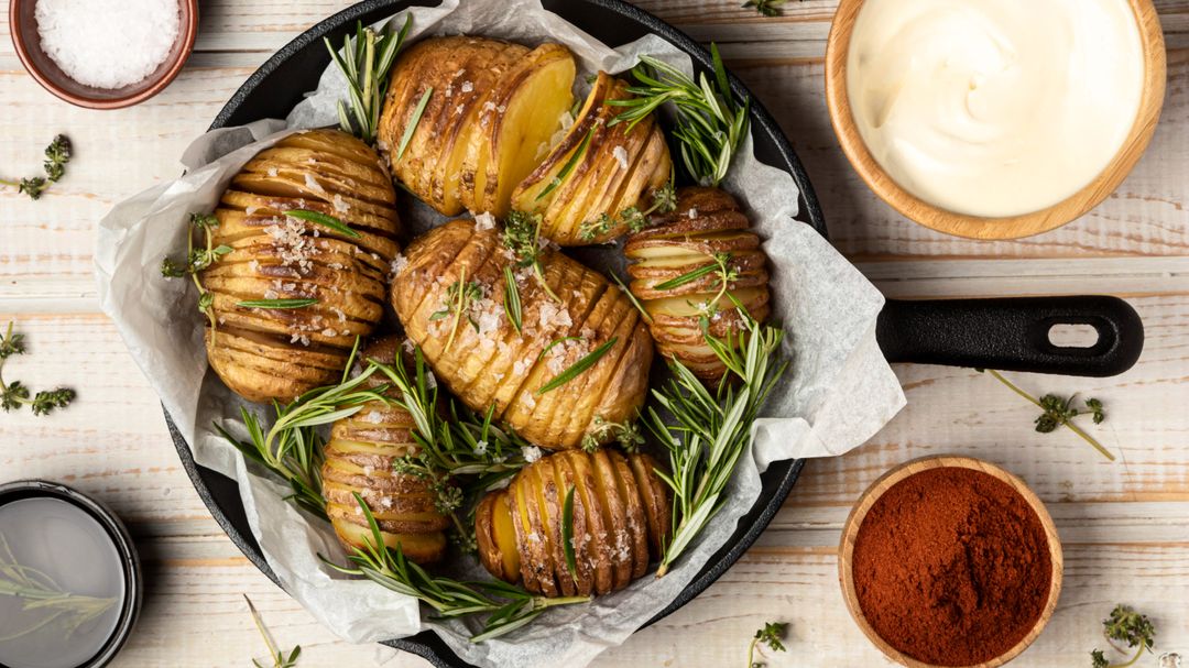 Draufsicht auf knusprige Fächerkartoffeln mit Rosmarin und grobem Meersalz in einer gusseisernen Pfanne, serviert mit cremigem Dip und Gewürzen auf hellem Holztisch.