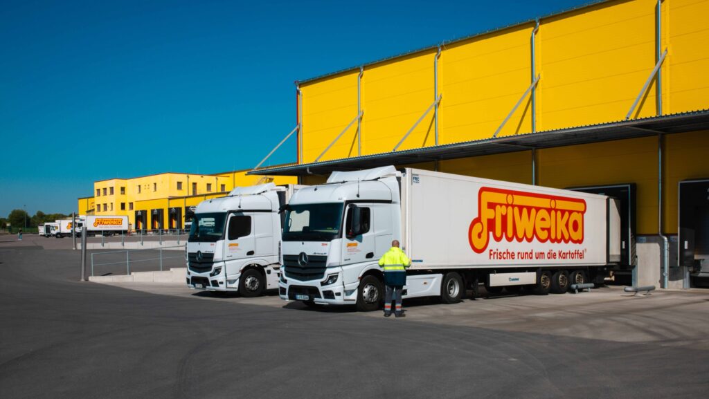 Zwei weiße Mercedes-Benz LKWs der Firma Friweika an einer gelben Logistikhalle bei der Beladung unter blauem Himmel.