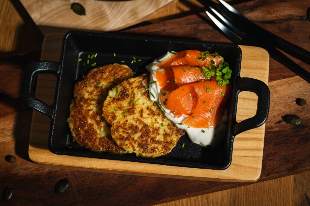 Serviervorschlag von Friweika: Knusprige goldbraune Kartoffelpuffer (Reibekuchen) serviert mit Räucherlachs, einem Klecks Sahnemeerrettich und frischen Kräutern in einer gusseisernen Pfanne.
