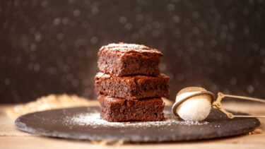 Drei gestapelte Schokoladen-Brownies auf einer dunklen Schieferplatte, bestäubt mit Puderzucker neben einem Teesieb.