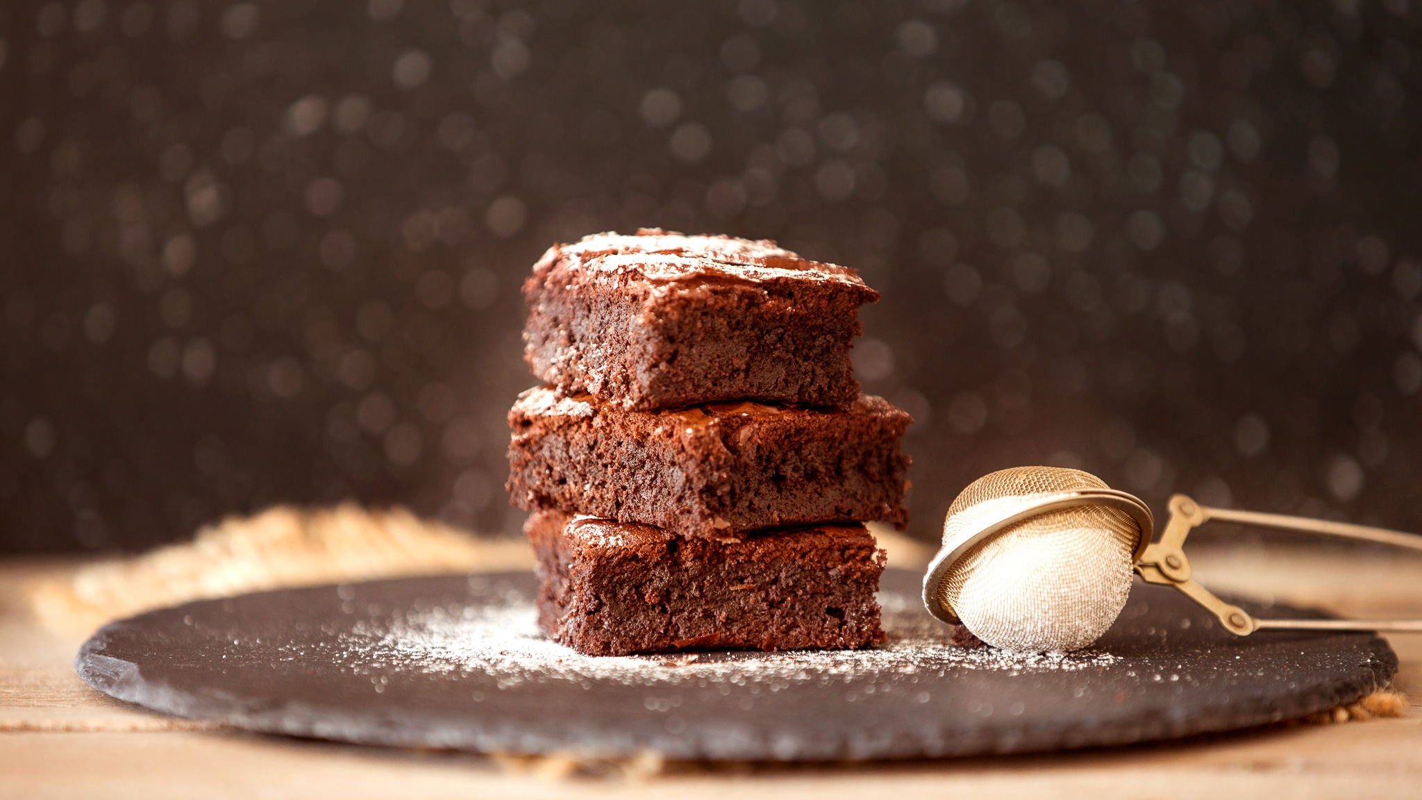 Drei gestapelte Schokoladen-Brownies auf einer dunklen Schieferplatte, bestäubt mit Puderzucker neben einem Teesieb.