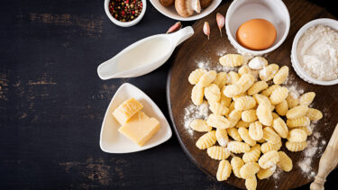 Draufsicht auf selbstgemachte Gnocchi auf einem Holzbrett mit frischen Zutaten wie Ei, Mehl, Milch, Pilzen, Knoblauch und Parmesan. Darunter steht der Text „Gnocchi - Zubereitungszeit: 60 Min. - Zutaten für 2 Personen - Leicht & lecker“.