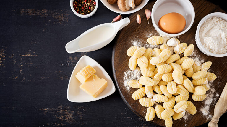 Draufsicht auf selbstgemachte Gnocchi auf einem Holzbrett mit frischen Zutaten wie Ei, Mehl, Milch, Pilzen, Knoblauch und Parmesan. Darunter steht der Text „Gnocchi - Zubereitungszeit: 60 Min. - Zutaten für 2 Personen - Leicht & lecker“.