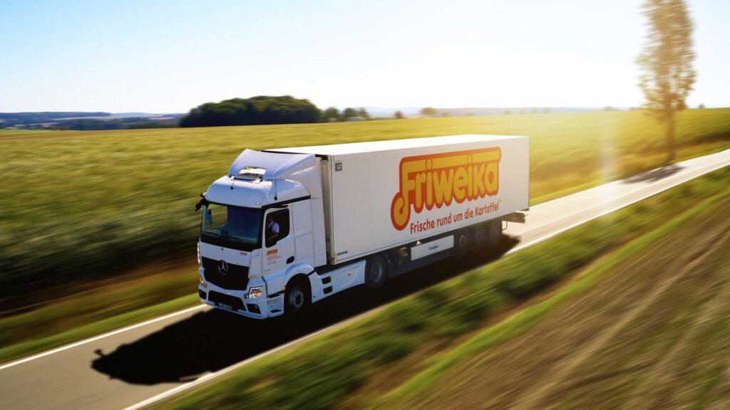 Weißer Friweika-Lkw fährt auf einer Landstraße vorbei an grünen Feldern.