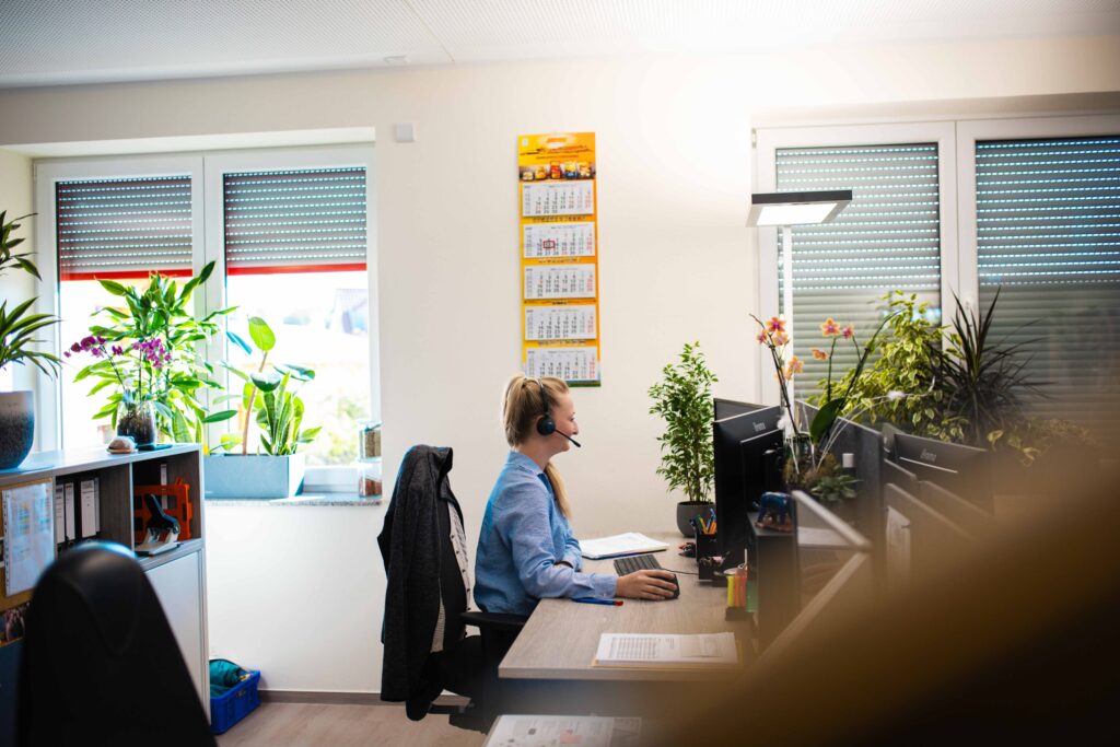Mitarbeiterin mit Headset arbeitet am Schreibtisch im modernen Büro mit Zimmerpflanzen.