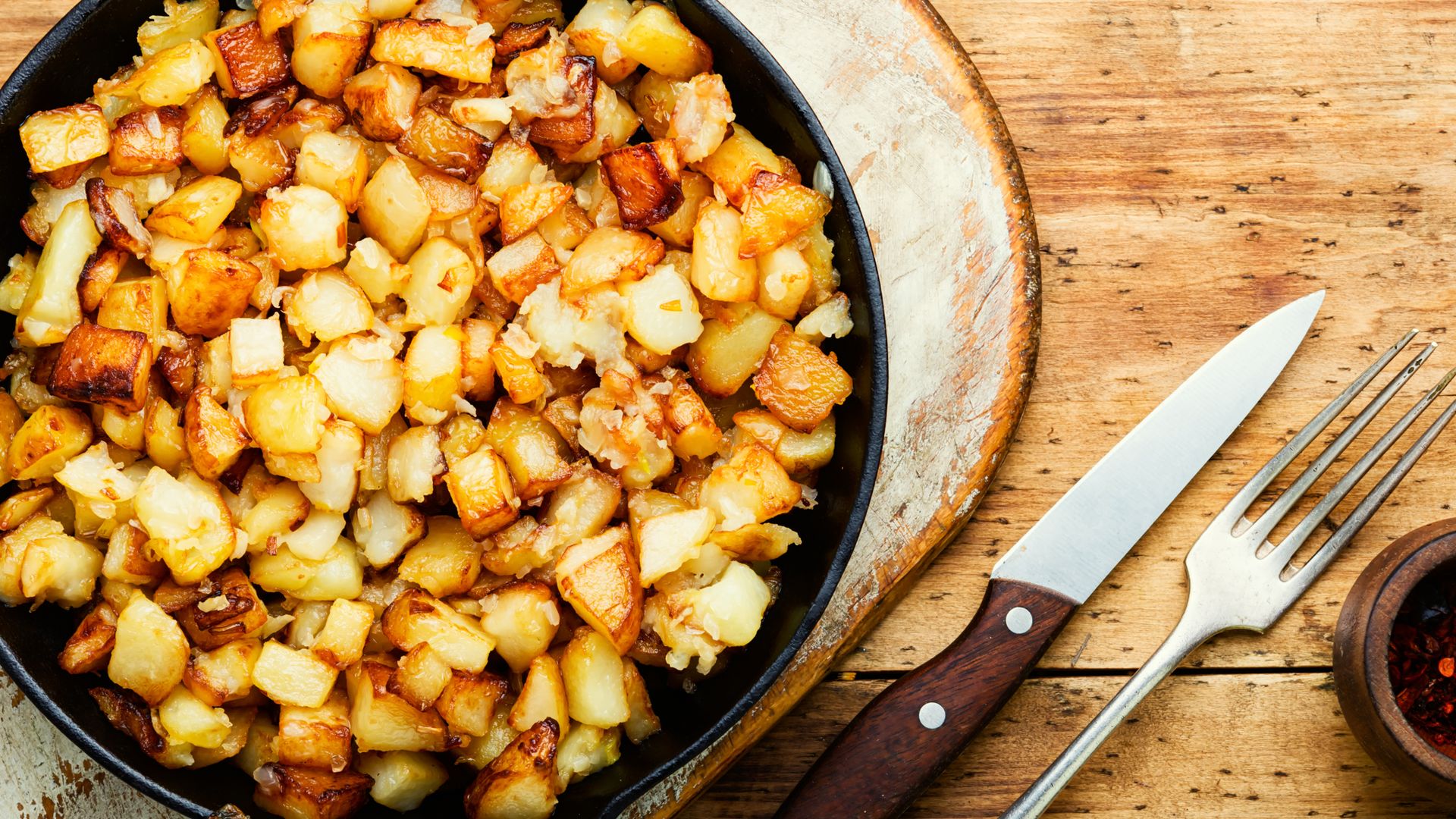 Nahaufnahme eines köstlichen, goldbraun gebratenen Kartoffel-Apfel-Rösti in einer schwarzen Gusseisenpfanne, platziert auf einem rustikalen Holzbrett. Daneben liegen ein scharfes Messer und eine Silber-Gabel. Ein traditionelles, wärmendes Winter-Pfannengericht.