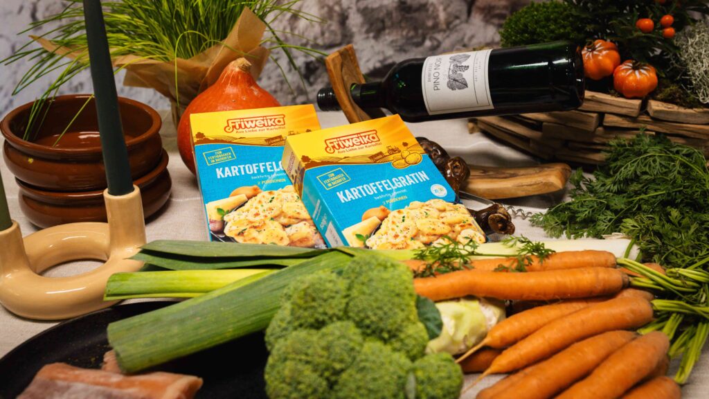 Zwei gelbe Friweika Kartoffelgratin-Packungen umgeben von frischem Gemüse wie Karotten, Lauch und Brokkoli sowie einer Weinflasche.