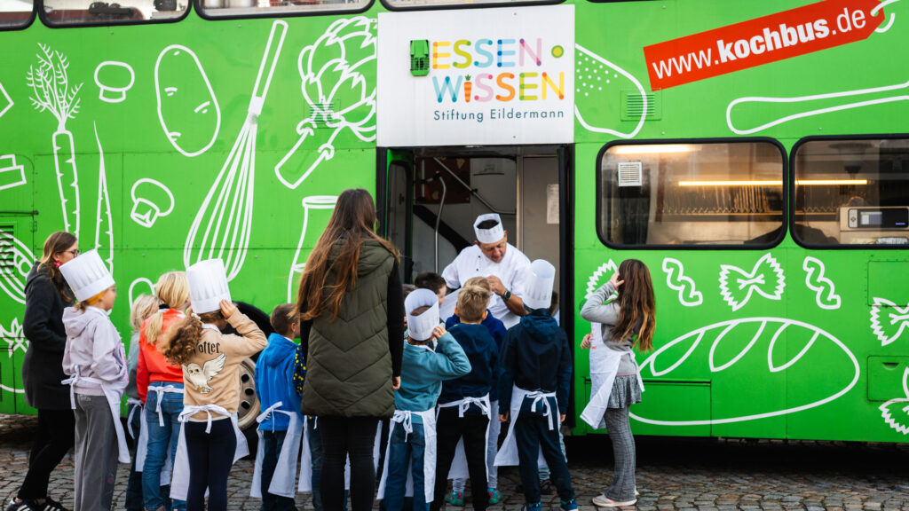 Kinder mit Kochmützen vor dem grünen Kochbus der Stiftung Eildermann