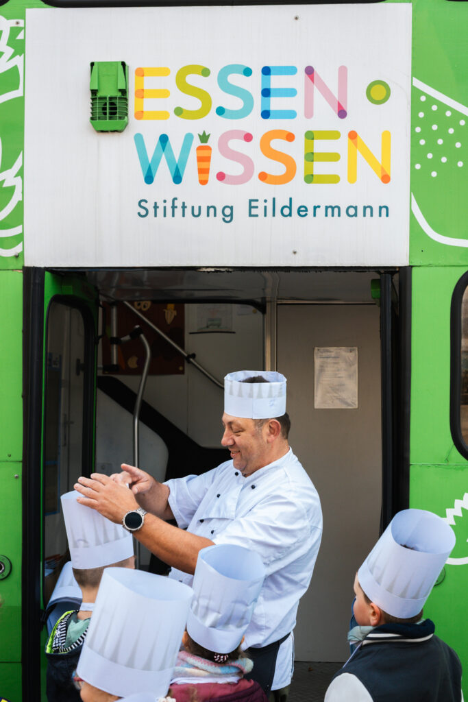 Koch setzt Kindern vor dem Eingang des Essen-Wissen-Kochbusses Kochmützen auf