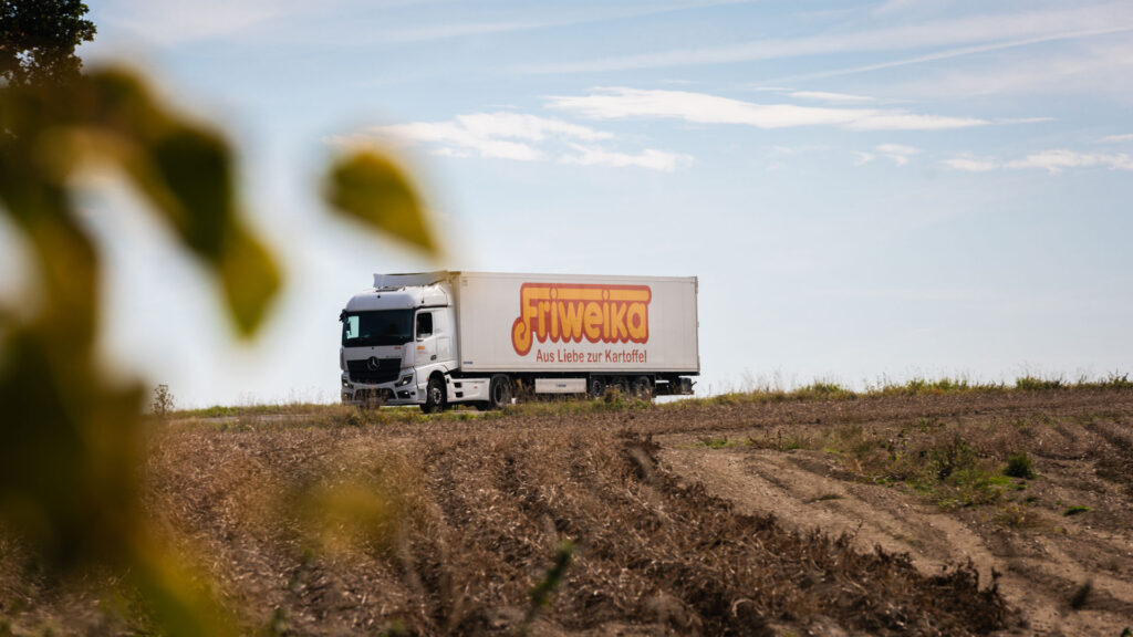 Weißer Friweika LKW mit der Aufschrift Aus Liebe zur Kartoffel fährt auf einer Landstraße am Feld entlang