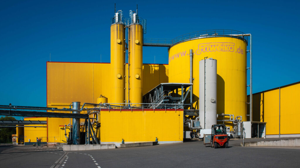 Das Foto zeigt ein Betriebsgelände bei hellem Tageslicht und klarem, blauem Himmel. Im Zentrum steht eine moderne Industrieanlage mit leuchtend gelben Gebäudewänden und zwei großen, gelben Silotürmen.